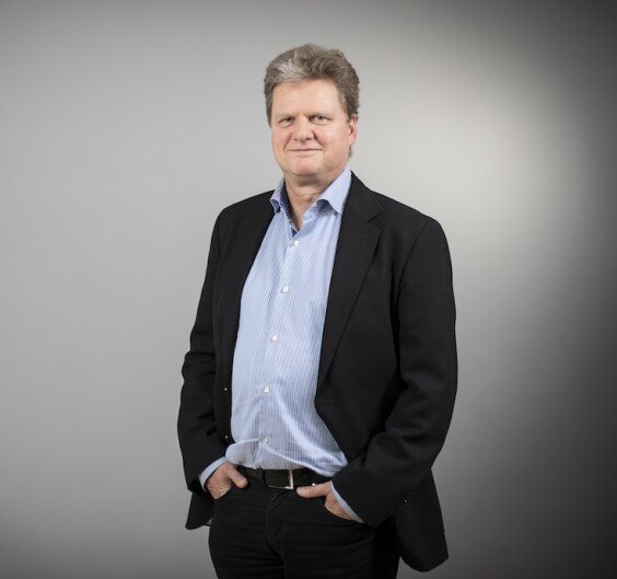 A white man with brown-grey hair. He is wearing a suit and a blue and white striped shirt. His gaze is neutral but friendly.