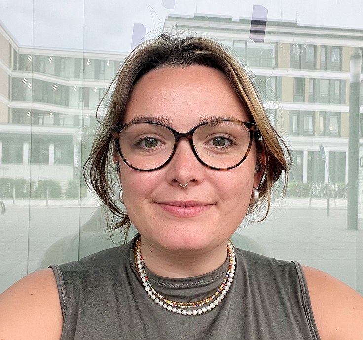 White woman with septum piercing, glasses and brown eyes smiles friendly into the camera.