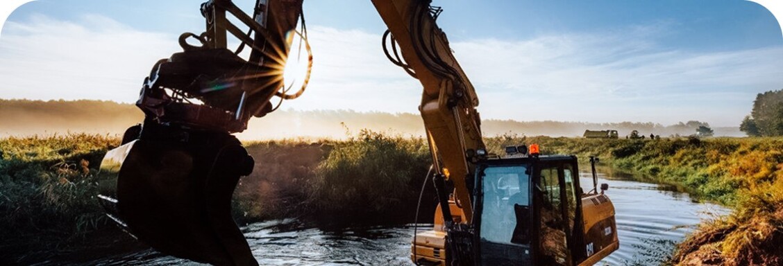 An excavator stands in a waterhole. In the background you can see a fog-covered field, which contrasts with the bright blue sky.