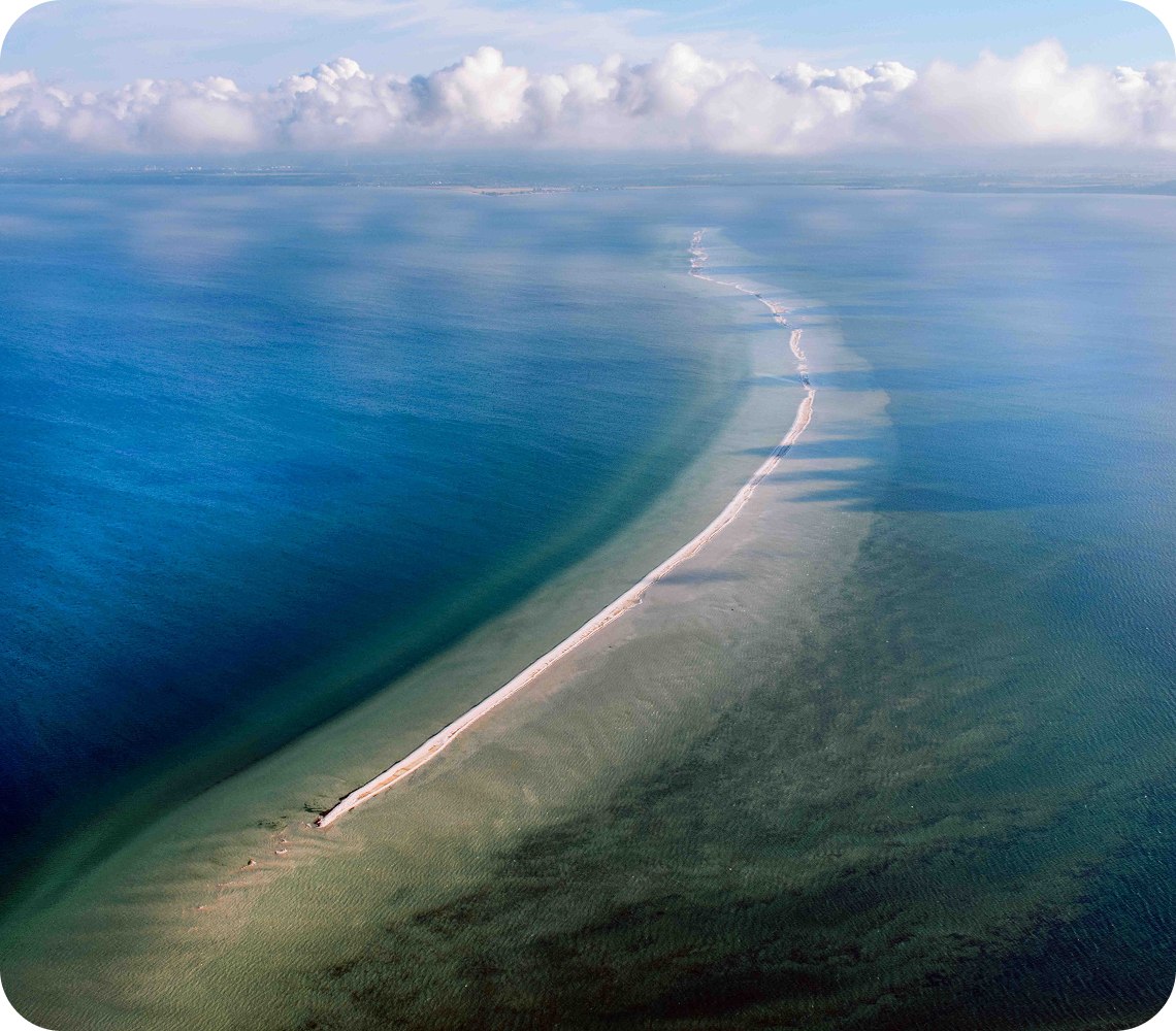 A very long sandbank stretches from top to bottom of the picture. Around it is dark blue water and above it white clouds.